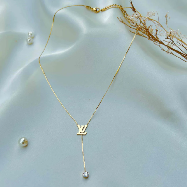 A gold-colored designer chain choker necklace with a pendant, displayed on a silky white background with decorative elements.